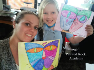 Mother and young daughter hold up matching art drawings from online class, labeled Painted Rock Academy 2020