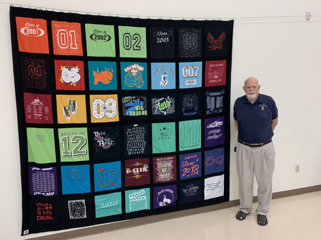 Older male teacher with gray beard stands by quilt constructed from blocks made out of many school t-shirts